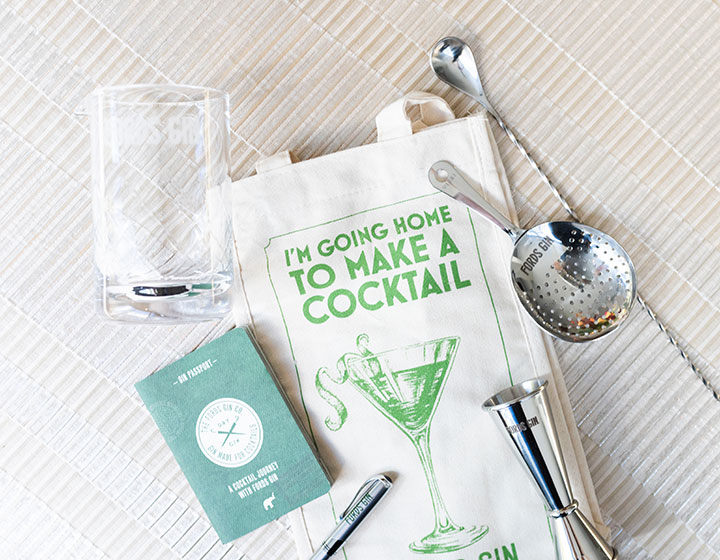 An overhead look at a cocktail kit: a bag, a glass, a jigger, a strainer, a notebook and a pen. 