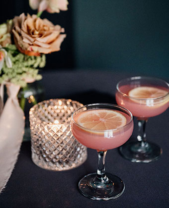 Close up of two pink cocktails, delicate flowers in a vase and a candle in the background